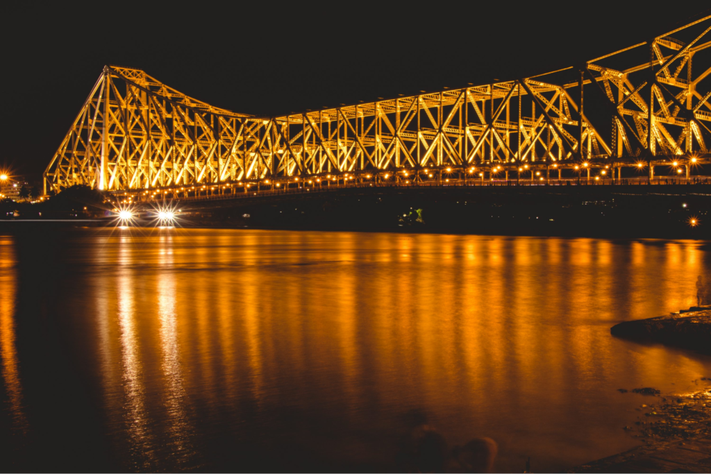 The image portrays the night view of Howrah Bridge with the shady Ganges below it