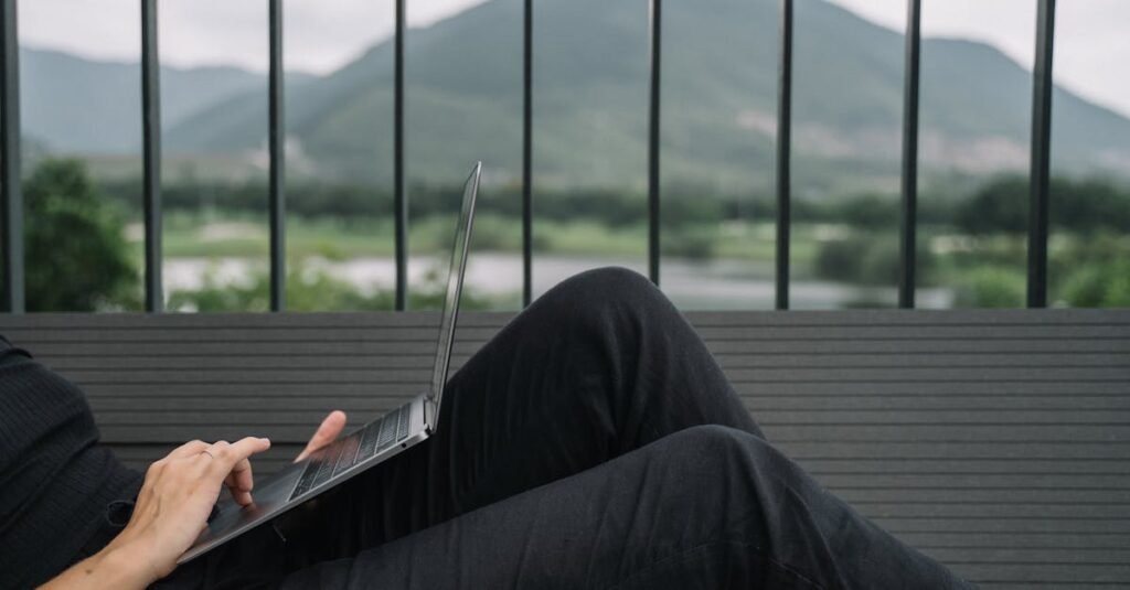 Person working remotely on a laptop, enjoying a serene mountain view from a balcony.